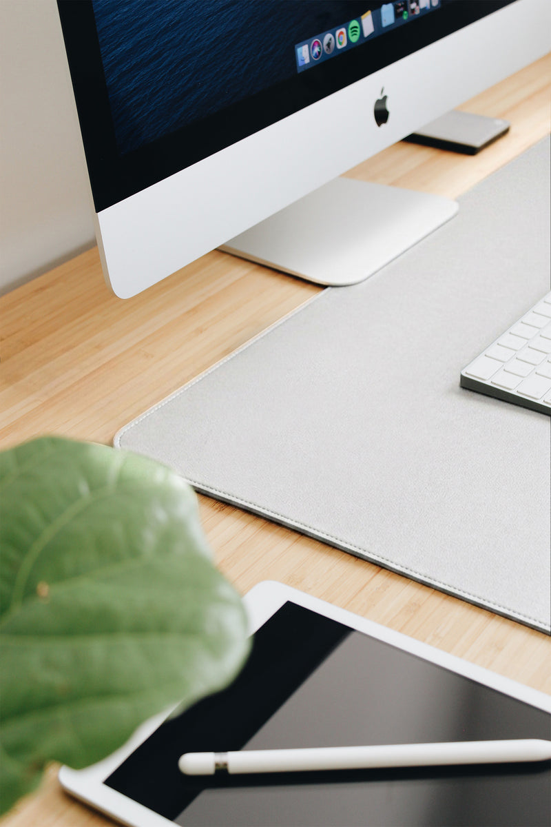 Close-up of an iMac and a white keyboard resting on a grey leather desk mat next to an iPad with a stylus.